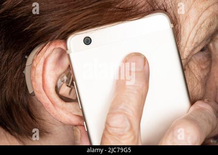 Elderly senior woman holding mobile phone next to her ear with hearing aid device, closeup detail Stock Photo