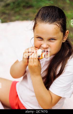 Portrait of little hungry girl wth dark hair holding food in hands, biting, sitting in park forest among green trees. Stock Photo