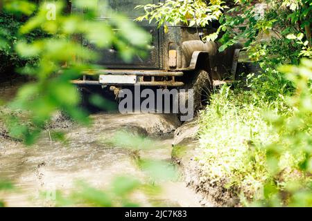 Back view of green russian off-road utility vehicle UAZ Hunter going up dirty road crossing river in forest among trees. Stock Photo