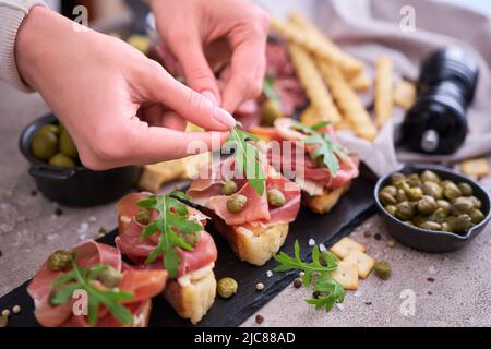 woman puts arugula leaves to prosciutto ham bruschetta with traditional antipasto meat plate on background Stock Photo
