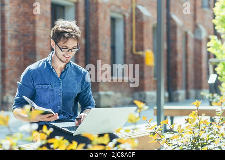 Young handsome creative young male architect with glasses sitting on the street on a bench with a laptop. works on the project, makes notes, drawings Stock Photo