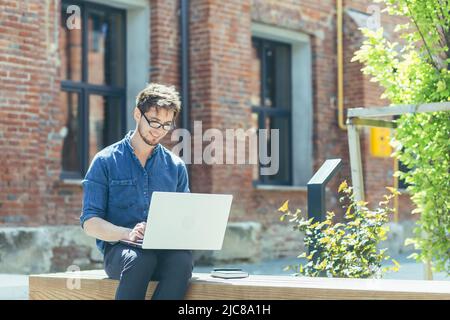 Portrait of a young handsome student in glasses and denim. He works online on a laptop, focused on sitting on a bench near the office center. Stock Photo