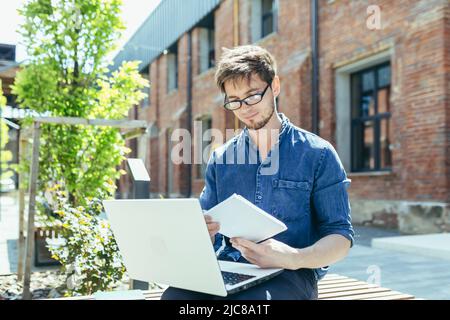 Young handsome creative young male architect with glasses sitting on the street on a bench with a laptop. works on the project, makes notes, drawings Stock Photo