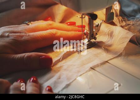 Photo of woman's hands with red nail polish in process of sewing linen dress using automatic sewing machine at home Stock Photo