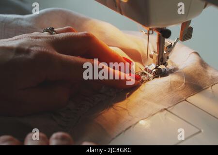 Photo of woman's hands with red nail polish in process of sewing linen dress using automatic sewing machine at home Stock Photo