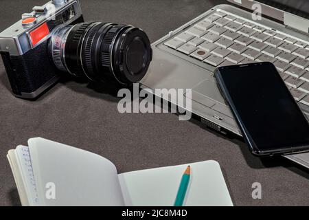 Top view of a photo camera, a laptop, a blank notepad with a green pencil and a cell phone, on a gray table. Copy space. Stock Photo