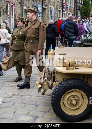 Haworth 1940's weekend (male dressed as soldier passing old Jeep, female in khaki, crowds on busy scenic Main Street) - West Yorkshire, England, UK. Stock Photo