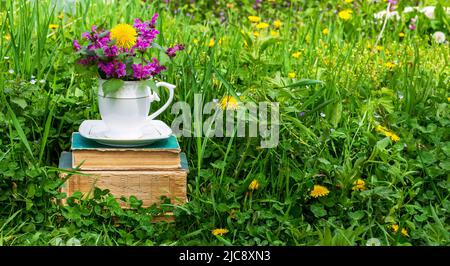 a white teacup with a bouquet of wildflowers on a stack of old books in a meadow with green grass on a sunny spring or summer morning Stock Photo