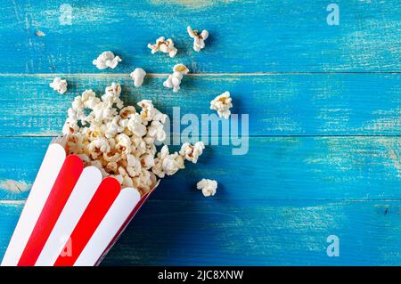 Fresh popcorn spilled from a cardboard striped cup on a blue wooden background with copy space. Top view. Stock Photo