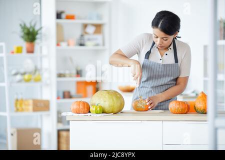 Description: Concentrated young black woman in stripped apron cutting pumpkin on board while carving pumpkin for Halloween in kitchen Stock Photo