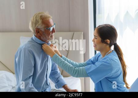 Young Asian woman nurse helping get dress to disabled elderly man in bed at retirement home. Millennial caregiver assisting handicapped senior patient Stock Photo