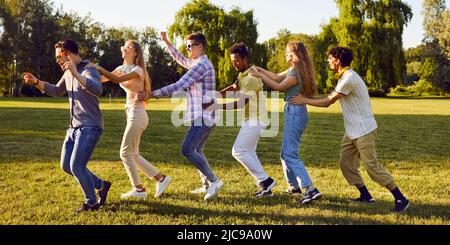 Cheerful active men and women having fun running in row one after another during gathering in park. Stock Photo