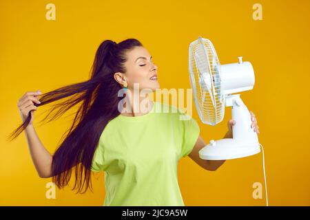Woman enjoys fresh air and escapes summer heat with electric fan isolated on orange background. Stock Photo