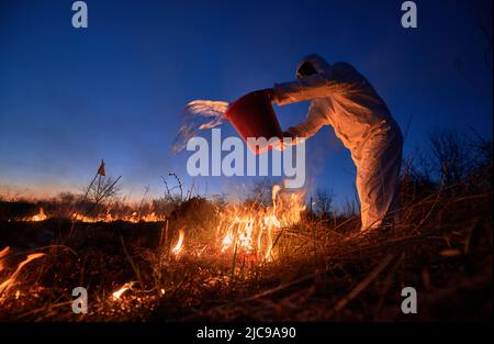 Research scientist fighting fire in field with blue night sky on background. Man in protective radiation suit and gas mask holding bucket and pouring water on burning grass with smoke. Stock Photo