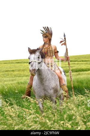 A young Indian girl is seen riding her pony in the prarie.  She is dressed up as a native American Indian and holds a spear in her hand. Stock Photo