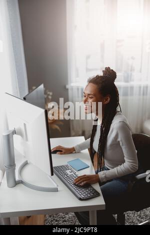 Smiling beautiful mixed-race female freelancer working on computer looking on screen at home Stock Photo