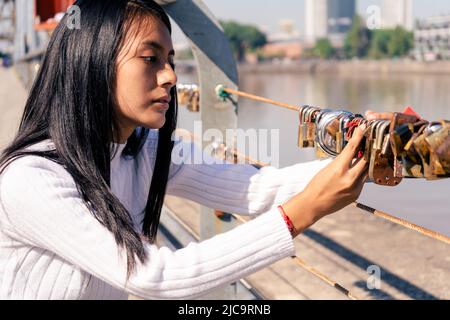 Beautiful black-haired Latin woman putting a lock on one of the bridges in a city. Concept fidelity, romantic love. Stock Photo