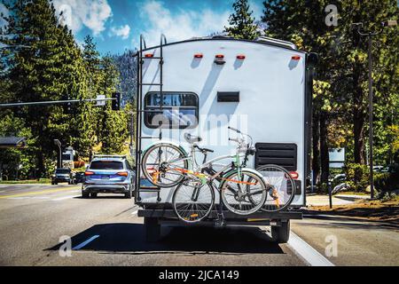 Camper with two bicycles tied to back on tree-lined highway outside of Lake Tahoe with mountains in distance on sunny summer da Stock Photo