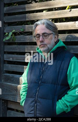 Mature man with grey hair and glasses leaning on the wooden fence, thoughtfully looking down with his hands in his pockets. Stock Photo