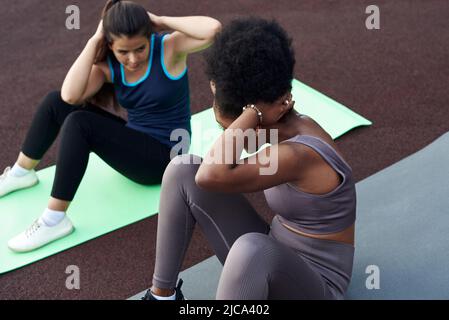 Cheerful young diverse female friends doing ab crunches on stadium in the park - friends together doing exercise outdoors Stock Photo
