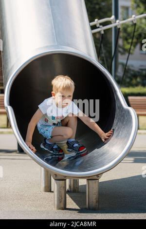 Joyful kid sliding in tube slide on playground. Kindergarten for elementary age children. Stock Photo
