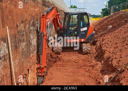 Worker Uses an Excavator to Dig a Trench Underneath and Road Next to a Reinforced Concrete Retaining Wall Stock Photo