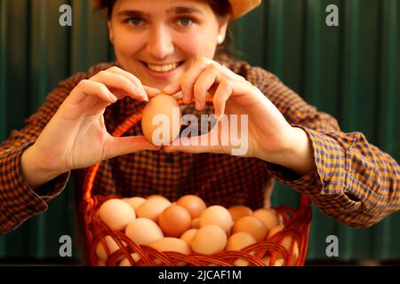 Many eggs in basket. Portrait of smiling young farmer in hat holding one brown organic egg on modern green background. Poultry farm. Eco agriculture, Stock Photo