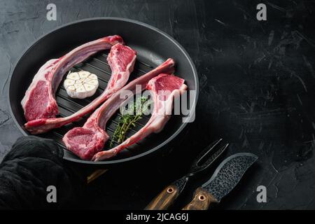 Organic pieces of mutton meat, Rack of lamb , raw with bone, with ingredients carrot orange, herbs, on frying cast iron pan, on black stone background Stock Photo