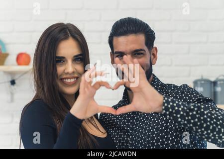 Interracial happy beautiful young couple folding their hands into a heart shape, smiling and looking at camera, standing in a kitchen. High quality photo Stock Photo