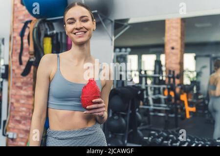 Excited young white slim woman standing in a well-equipped gym, smiling at camera, holding a red fake human heart. High quality photo