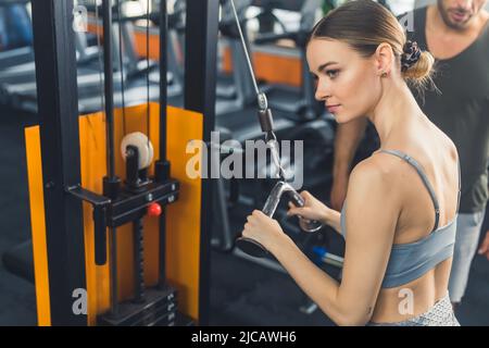 Caucasian brunette athletic woman in her 20s with her hair tied up in a bun, focusing on doing an exercise for her triceps and back muscles. Personal male trainer in the background. High quality photo Stock Photo