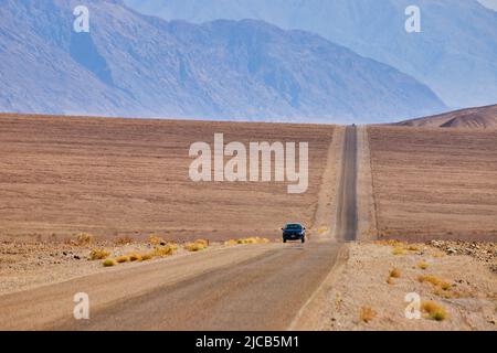 Long straight road with car driving through empty desert plains and hills by mountains Stock Photo
