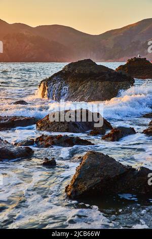 Ocean waves crashing and pouring over boulders by mountains Stock Photo