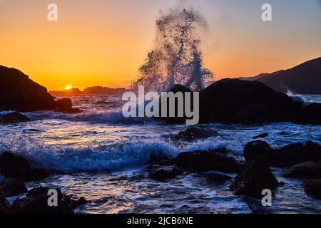 Large wave crashing into rocks on beach during gorgeous sunset Stock Photo