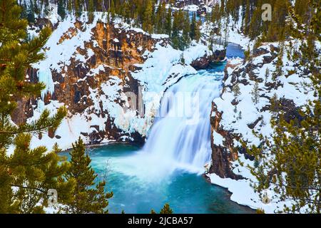 Pine trees frame snowy cliffs and stunning blue waterfalls in Yellowstone Stock Photo