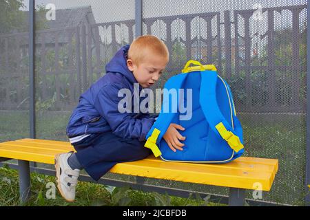 funny boy sitting on a bench with a backpack and waiting for the bus Stock Photo