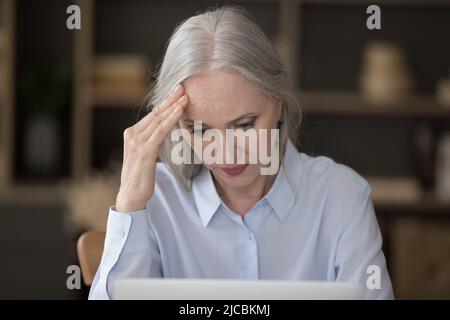 Serious mature businesswoman looks at laptop touch head feels stressed Stock Photo