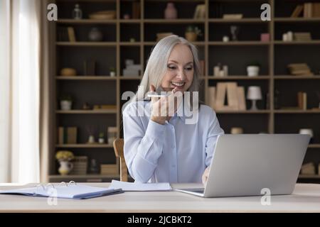 Middle-aged businesslady talks on speakerphone seated at desk with laptop Stock Photo