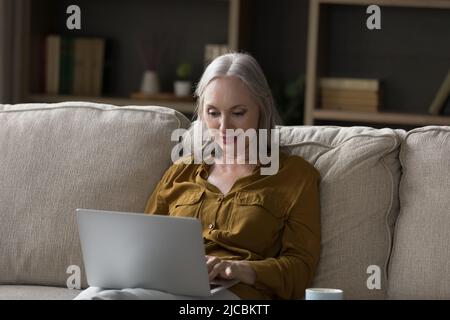 Pretty older woman sit on sofa using laptop Stock Photo