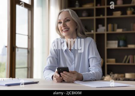 Older woman sit at desk with smartphone looking into distance Stock Photo