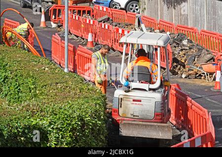 Broadband super fast coil of orange fibre optic cable workmen place it in trench hole dug by mini excavator in pavement rural Essex village England UK Stock Photo