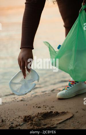 Cropped photo of African woman collecting spilled garbage from sand on beach in green plastic bag. Ecology, pollution. Stock Photo