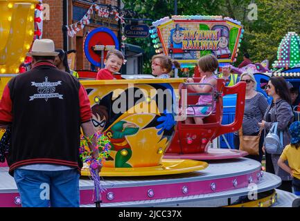 Children enjoy riding in a large teacup street ride while parents look on smiling at St George’s Day Celebration 2022, Pinner, Harrow, London. Stock Photo