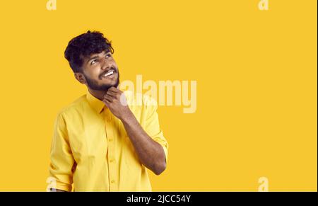 Cheerful stylish Indian guy pensively looks at copy space touching his chin on orange background. Stock Photo