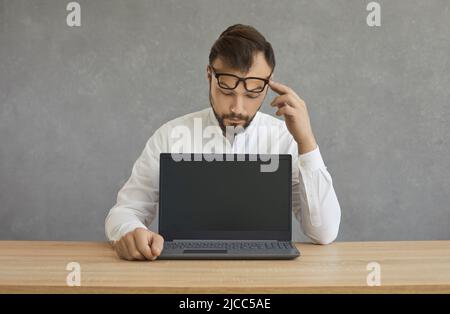 Serious man sitting at desk and looking at empty mock up screen of modern laptop Stock Photo
