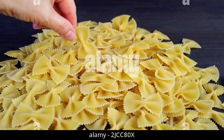 Hand Picking a Piece of Farfalle or Bow-tie Pasta from Its Pile on Black Wooden Background Stock Photo