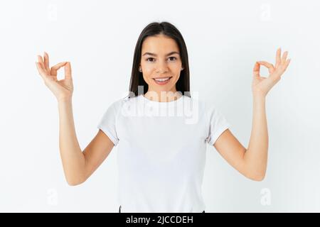 Portrait of happy serene young woman meditating wearing plain white t-shirt raising hands with lotus gesture, standing in white studio Stock Photo