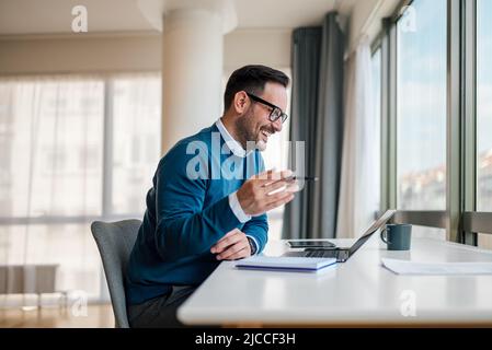 Smiling young professional working on laptop while sitting at desk. Happy businessman is analyzing successful strategy. He is wearing smart casuals in Stock Photo