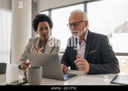 Female executive discussing with senior professional over laptop. Business colleagues are planning at desk. They are wearing formals in office. Stock Photo