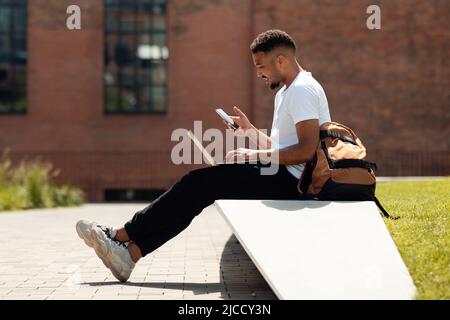 Modern black man with laptop and cellphone sitting outdoors and surfing internet or texting, side view, free space Stock Photo